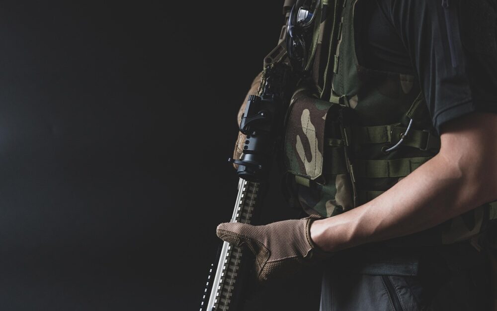 Cropped image of a white man in fatigues holding a gun pointed downwards.